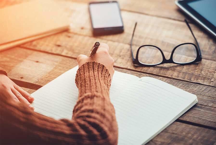 A tabletop and a person’s hand holding a pen and starting to write in a notebook.