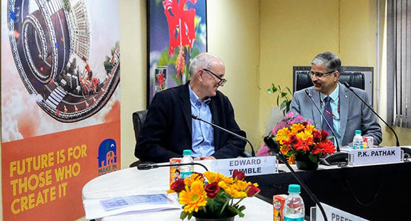 Two men seated at a conference table during a meeting about cancer research priorities with the Ministry of AYUSH. On the left is Edward Trimble wearing glasses and a dark jacket with a nameplate in front of him. On the right is P.K. Pathak in a gray suit speaking into a microphone.