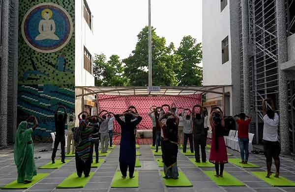Group yoga session at the Delhi State Cancer Institute. Several people are practicing yoga on bright green mats arranged in rows between buildings. Participants include patients and possibly staff in various colored clothing, performing stretches with arms raised.