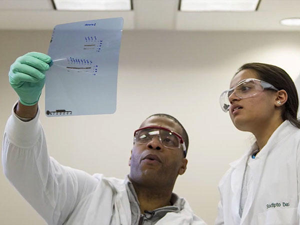 A male and a female researcher look at a projector sheet the male researcher is holding up. Both wear white lab coats and safety goggles; the male researcher also wears a green latex glove. Ceiling lights of an office environment are in the background.