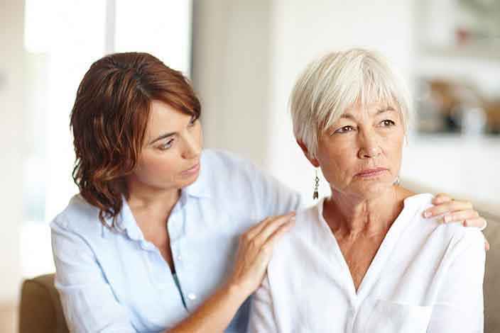 A woman comforting an older woman, gently placing a hand on her shoulder. Both are seated and appear thoughtful. The younger woman looks at the older woman with empathy and concern, while the older woman gazes into the distance. 