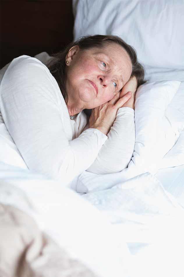 An older woman lying in bed, resting her head on her hands. She has a thoughtful and somewhat melancholic expression. 