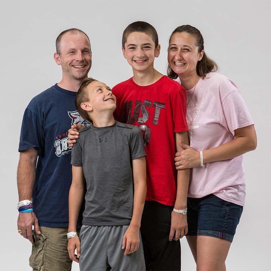 A joyful family of four, including two parents and two young boys, stands together smiling brightly.