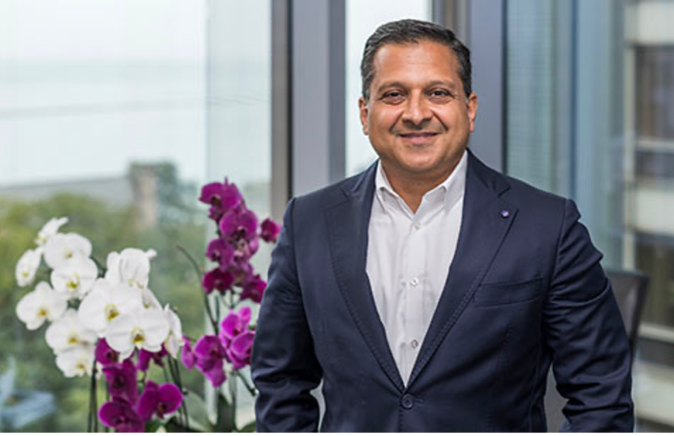 Ali Shilatifard wears a navy suit and white shirt is smiling while standing in an office with large windows. The background shows a blurred view of trees and buildings, and there are white and purple orchids on the left side.