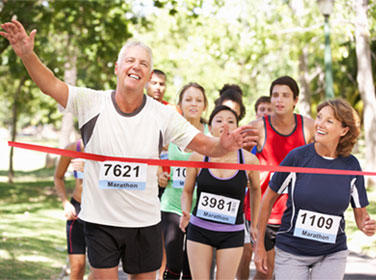 Group of runners of various ages crossing a finish line during a race, with an older male runner with race number 7621 celebrating with his arms raised while surrounded by other happy participants wearing racing bibs.