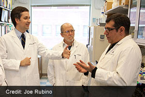 (From left to right) Victor Velculescu, M.D., Ph.D., Bert Vogelstein, M.D., and Nickolas Papadopoulos, Ph.D. in white lab coats examining documents in a laboratory setting at Johns Hopkins University.