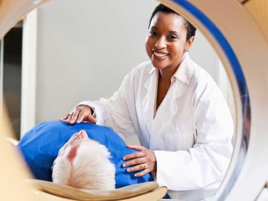 A healthcare professional assists an elderly patient during a medical scan. The patient is lying on their back, partially inside a circular imaging machine, while the professional, dressed in a white coat, offers reassurance with a smile. 