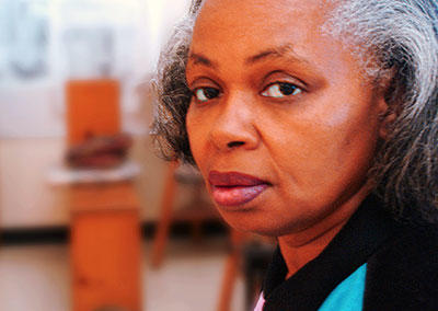 An elderly Black woman with white, grey and black hair looks at the camera. She has a neutral expression. The background has some furniture, indicating she is indoors.