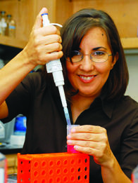 A smiling woman with dark hair wearing glasses and a dark blouse looks at the camera and smiles while holding a pipette above a sample array.