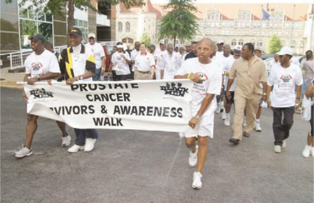 A large group of men, dressed in casual clothing, walk through the streets. Three men at the front hold a banner that reads “Prostate Cancer Survivors & Awareness Walk”. The crowd is predominantly Black. 