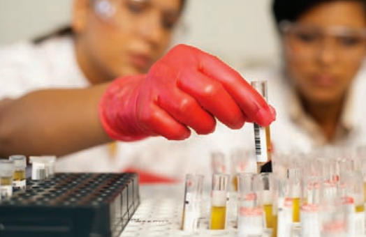 Two women, wearing safety goggles and lab coats, look at samples in various vials. The women’s faces are blurred in the background, due to depth of field, while one researcher’s hand, wearing a red latex glove, is in focus while picking up a sample vial.