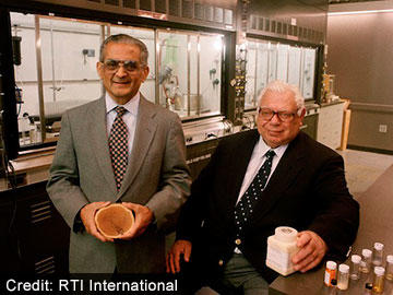 Mansukh Wani stands next to Monroe Wall who is seated in a lab environment. Both men wear suits and ties as well as glasses. Dr. Wani holds tree bark in his hands; Dr. Wall holds a large rectangular medicine bottle. The background is mostly grey and metallic.