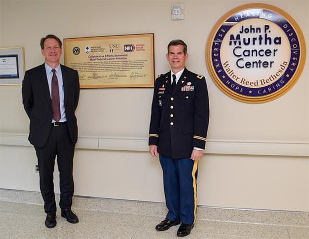 NCI Director Norman Sharpless, M.D. (left) standing with Colonel Craig Shriver, M.D.  in a hallway next to a sign for the John P. Murtha Cancer Center. The wall displays a circular emblem and a plaque with text about the center.