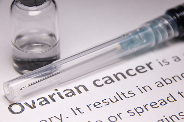 A close-up of a syringe and medical bottle containing clear liquid. These two items are sitting on a piece of paper that shows the definition of ovarian cancer. The words “ovarian cancer” are bold; the rest of the text is mostly cropped off the image, though what is shown indicates it is a definition of ovarian cancer.