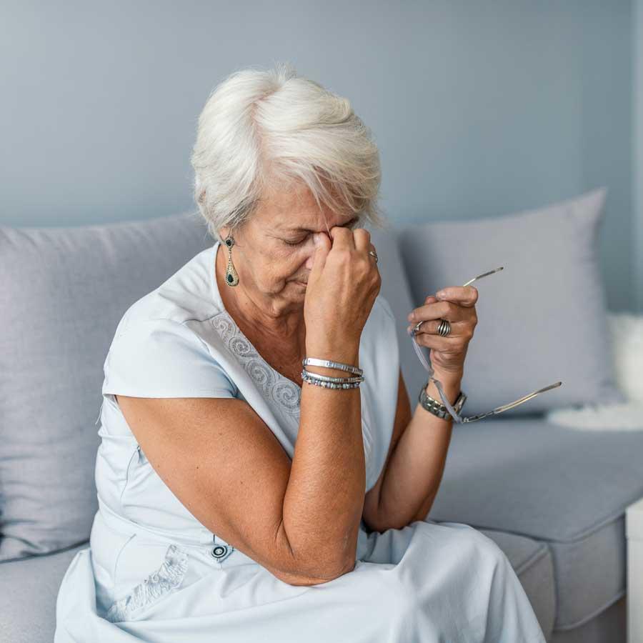 An elderly woman sits on a couch holding her glasses in one hand and rubbing her forehead with the other, appearing to be in discomfort.
