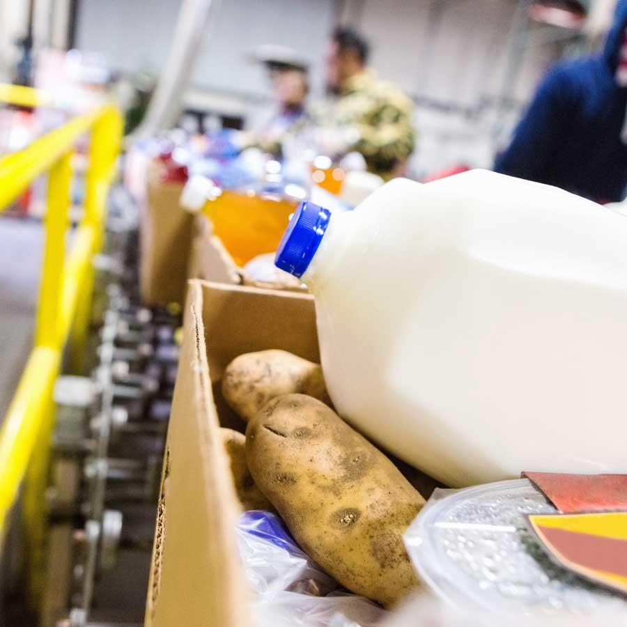 Food bank workers sorting through donated groceries, with a close-up view of potatoes and milk containers in cardboard boxes, while volunteers work in the background.