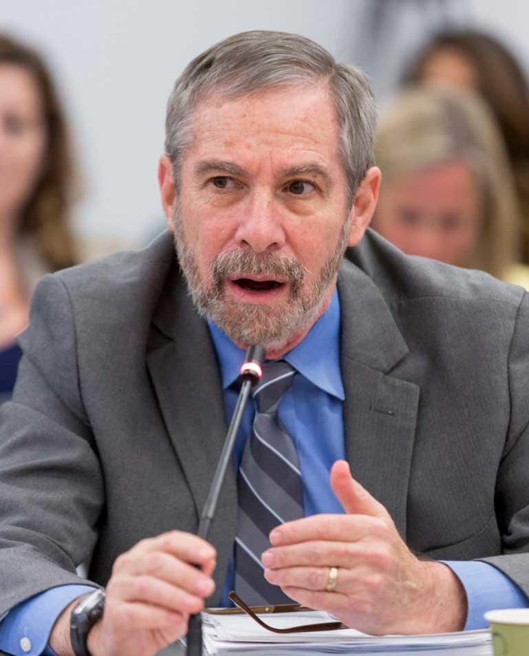 Director Douglas Lowy sitting at a table and speaking into a microphone at a meeting of the National Cancer Advisory Board. Dr. Lowy has short gray hair and beard and is wearing a grey suit, blue shirt and striped tie.