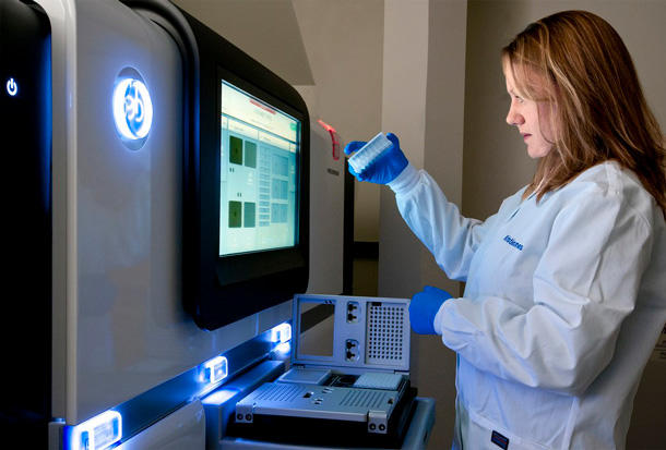 A female research technician holds up a small instrument in front of a large machine at the Advanced Technology Research Facility at NCI's Frederick National Laboratory for Cancer Research. The technician is wearing a white lab coat and blue gloves and has shoulder length brown hair with highlights. The large machine has a screen and lights on it; the instrument being held fits in to a smaller device that is below the screen.