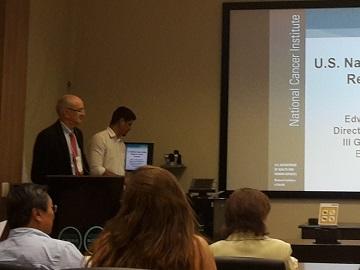 A view from the audience of a presentation at the Global Cancer Symposium. Dr. Ted Trimble stands at a podium in the front of the room. A projection screen behind them shows part of a slide titled 'U.S. National Cancer Institute' with additional text partially visible. Several attendees can be seen from behind in the foreground, watching the presentation