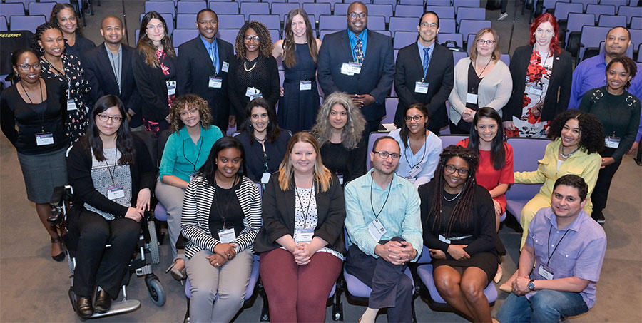 A group of people, all of whom are dressed in professional attire, look at the camera with happy expressions. Some are seated and some are standing; the chairs and surroundings indicate that this photo was taken in a theater. They all wear lanyards around their necks. 