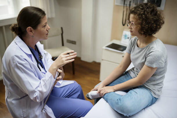 A female doctor and female patient are sitting and having a conversation. The doctor has dark hair tied back and is wearing a white coat with blue scrubs and a stethoscope around her neck. She is holding a clipboard with one hand and gesturing with the other. The patient has short curly dark hair and is wearing glasses, a grey t-shirt and blue jeans. Part of the examining room is visible in the background.