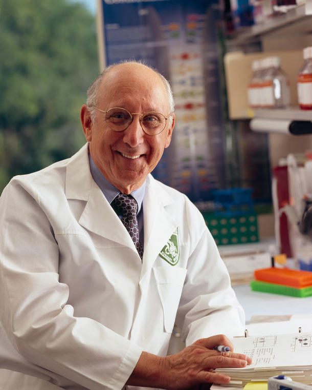 Stephen Rosenberg looks at the camera and smiles. He is an elderly balding man wearing glasses, a white coat with a crest on the chest, and a shirt and tie. He is holding a pen in one hand over an open book. His laboratory is visible in the background. 