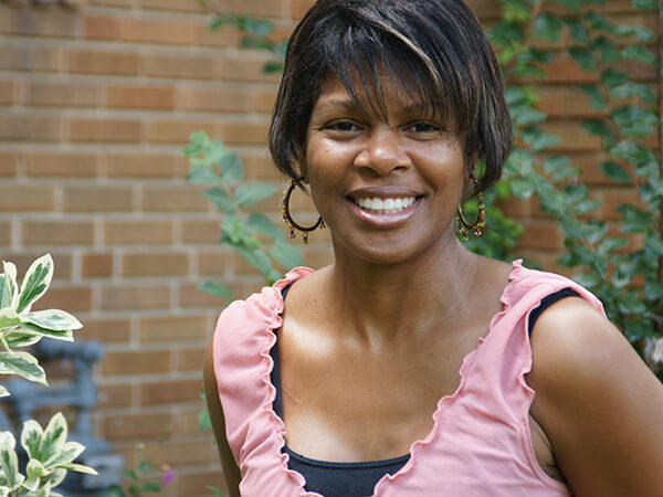 Portrait of Melanie Nix, smiling while wearing a pink ruffled top and earrings, photographed outdoors with greenery and brick in the background