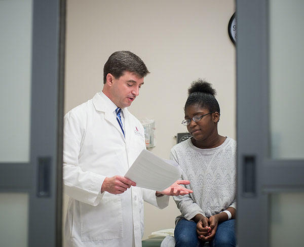 A doctor looks at a piece of paper and speaks to his patient. The doctor is a white middle-aged male, wearing a lab coat, button up shirt and tie, He is standing and gesturing as he speaks. The patient, a younger Black woman with glasses, a long sleeve grey sweater and jeans, sits on the exam table and looks slightly distressed. Out of focus doors to the room are in the foreground.