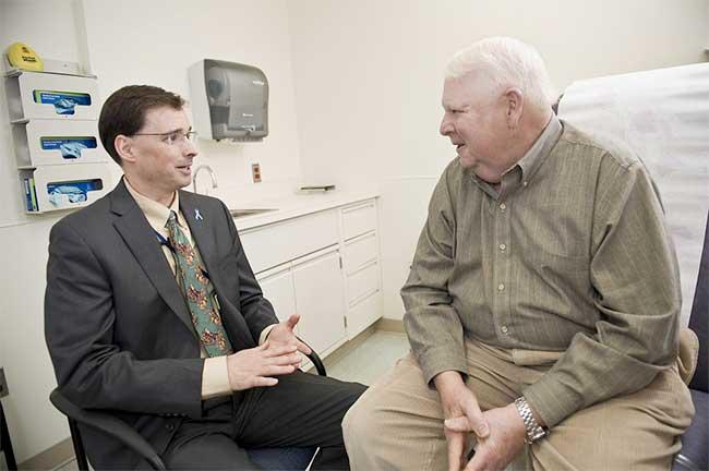 A doctor and a patient, both sitting, speak to each other. The doctor is middle aged with short dark hair, glasses, a grey suit and tan shirt with patterned tie. He’s sitting in a chair. The patient is an elderly man in a green button up shirt and khakis with white hair; he sits on an exam chair. A sink, cabinets, and gloves are in the background.