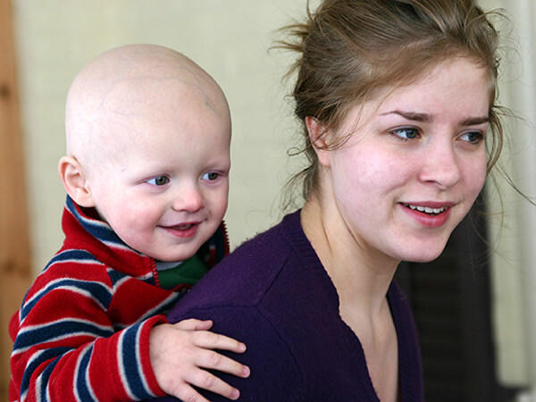 Child with a bald head, is smiling while playfully holding onto the woman’s shoulders. The woman looks content as she interacts with the child.
