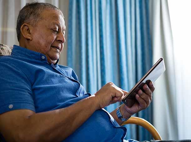 Elderly man in a blue shirt using a tablet or mobile device while seated in a chair near a window with blue curtains.