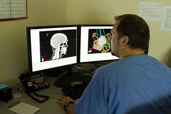 Medical professional in scrubs reviewing brain scan images on dual computer monitors, showing both traditional X-ray and enhanced colorized views.