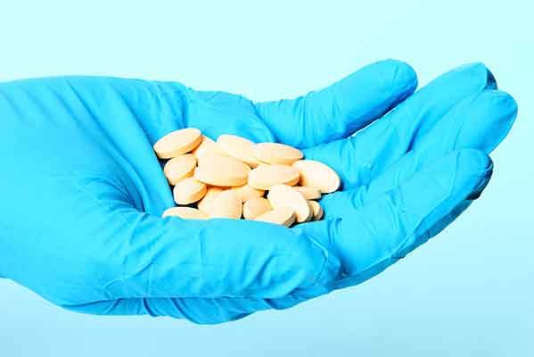 An outstretched hand wearing a blue latex glove holds a handful of oval pills which are light orange in color.