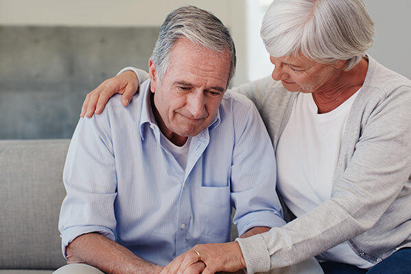 Elderly woman comforting a man by placing her hand on his shoulder as they sit together on a couch, with the man appearing distressed or contemplative.