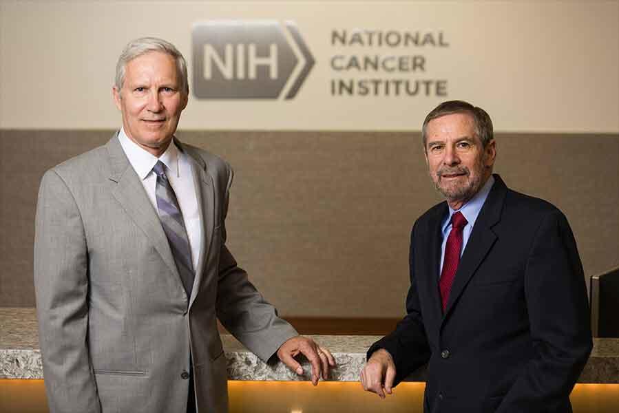 John T. Schiller, Ph.D. (left) and Douglas R. Lowy, M.D. (right) in business suits standing at a marble-topped table in front of the National Cancer Institute logo.
