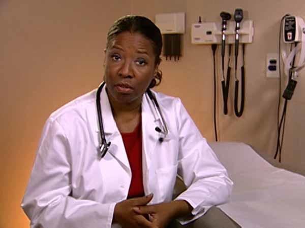 A Black female adult doctor, wearing a white lab coat, red shirt, and stethoscope around her neck, looks at the camera with a serious expression. She is seated next to an examination table. In the background, medical tools hang on the wall.