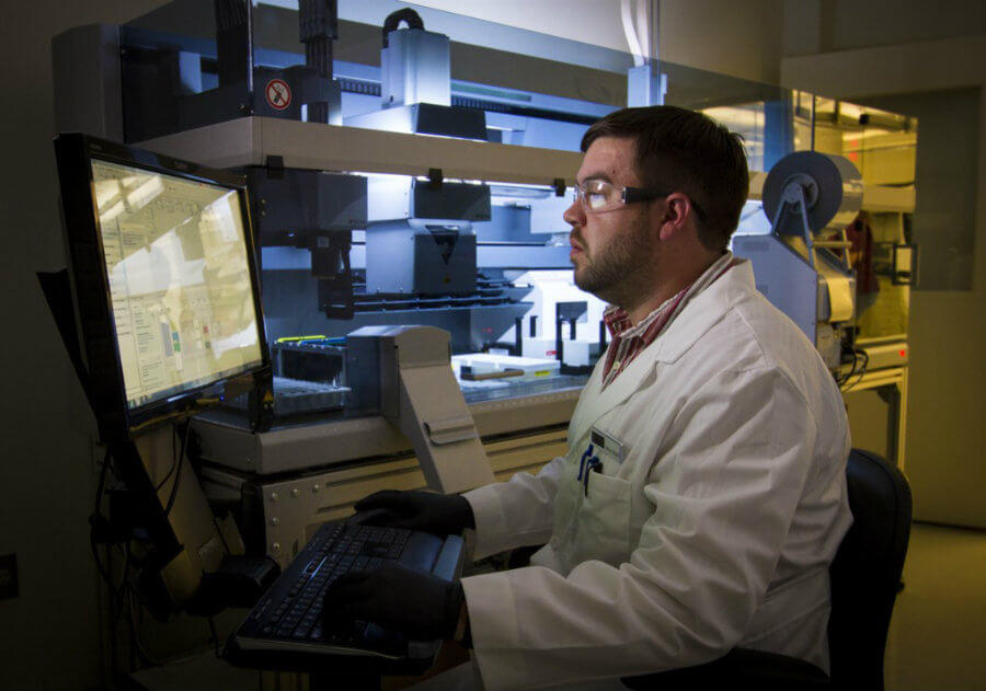 A scientist in a lab coat and safety glasses is working at a computer station in a laboratory. The lab is equipped with advanced machinery in the background. The scientist is focused on data displayed on the screen.