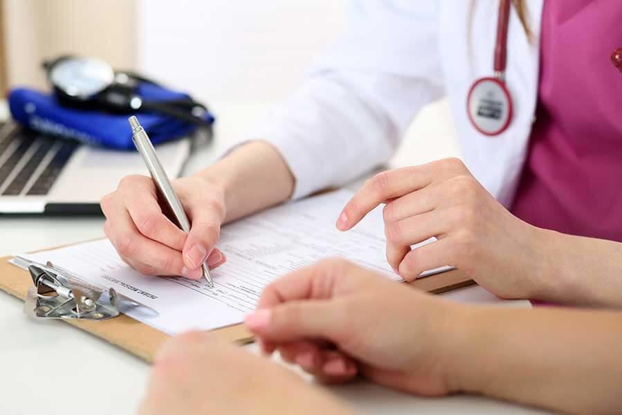 Medical professionals in white coats and a pink scrub top completing medical forms at a desk, with a patient's hands visible reaching toward the paperwork.