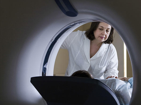 A female doctor looks at her patient as the patient enters a CT machine. The doctor has shoulder length brown hair and a white coat. The back of the headrest going into the CT machine is mostly in shadow.