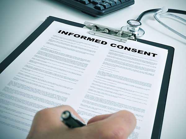 A close-up of a hand holding a pen to a clipboard holding a piece of paper. The words “Informed Consent”  are printed in large letters at the top of the paper, with smaller text in two columns below. A stethoscope and part of a keyboard are in the background.