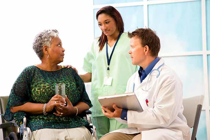 An older adult seated in a wheelchair, holding a glass, while interacting with a doctor and a nurse. The doctor, with a stethoscope and clipboard, is engaged in conversation, while the nurse offers support by gently placing a hand on the patient’s shoulder. 