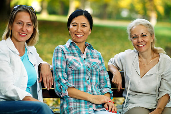 Three middle-aged women, all looking at the camera and smiling, sit on a park bench. They are dressed in casual clothing. The background is green and indicative of a park.