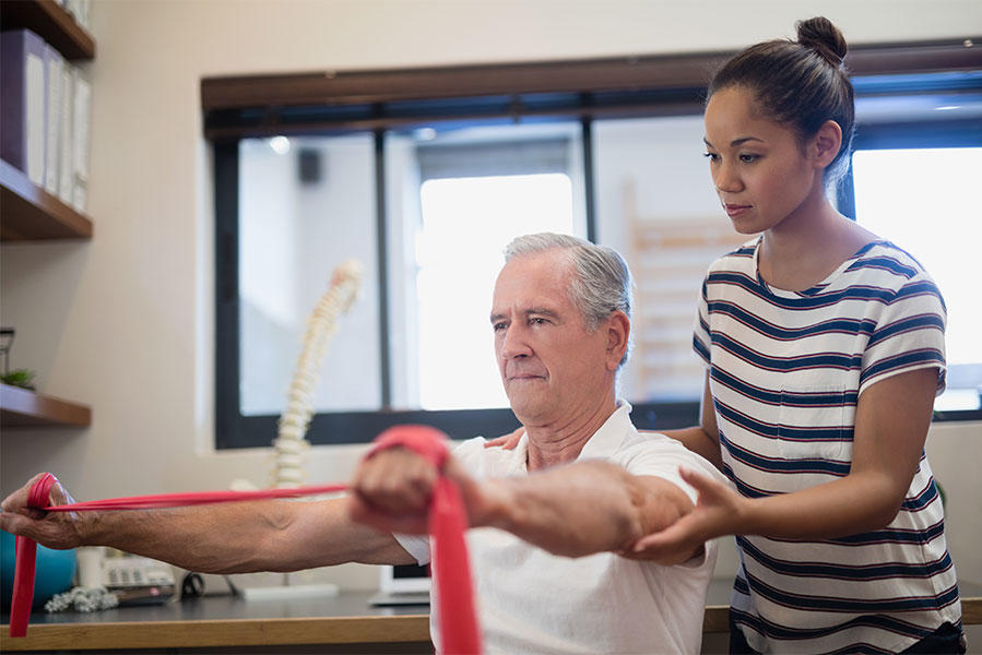 Physical therapist assisting an older male patient with resistance band exercises in a bright therapy clinic, with the patient stretching the red resistance band while the therapist provides guidance and support.