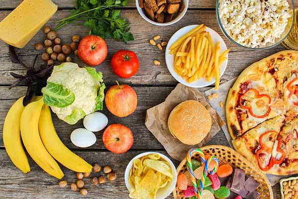 Overhead view of various foods arranged on wooden surface, including fresh produce like bananas, apples, and cauliflower on the left side, and processed foods like pizza, burgers, french fries, and snacks on the right side.