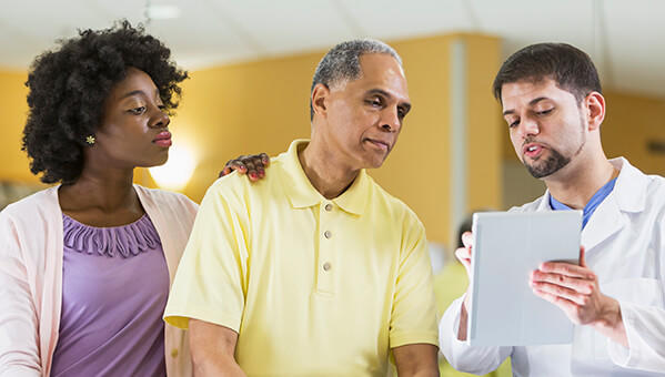 Healthcare provider in white coat explaining information on a tablet to a man in yellow shirt and woman in purple blouse in a medical setting.