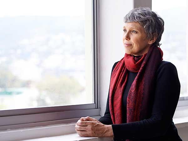A woman with short gray hair wearing a black top and red scarf gazes thoughtfully out a large window. She is standing with her hands clasped together.