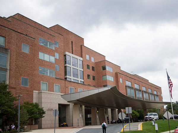 An exterior photo of The National Institutes of Health Clinical Center. It is approximately 10 stories high and mostly composed of brick, with a large, modern, curves steel awning in the front. An American flag can be seen in a patch of grass in front of the awning. Vehicles can be seen under the awning. The sky is grey and cloudy.