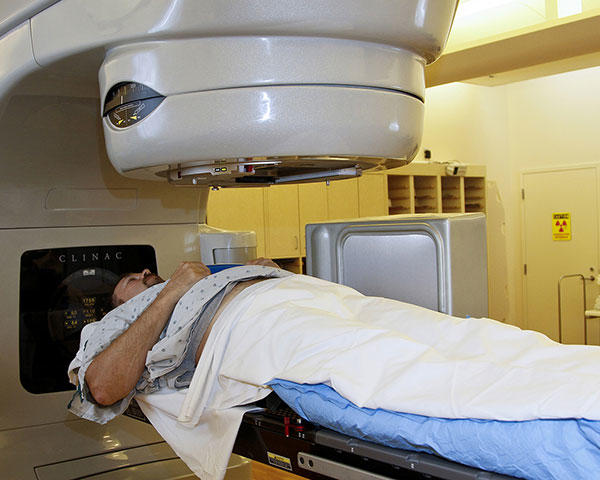 A patient lays on his back under a radiation machine. The patient is dressed in a hospital gown and covered with a blanket. The machine is large and grey with a cylindrical section directly above the patient’s chest. The background is tinted yellow and there are cabinets and a door.