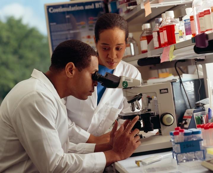 A male researcher looking through a microscope in a laboratory with a female researcher standing near.