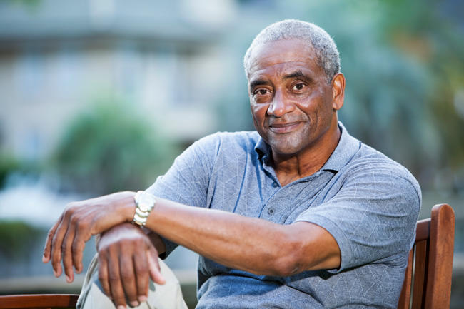 A middle-aged African American man looks at the camera with a happy expression. He has closely cropped grey hair and is wearing a grey short sleeve polo shirt and khakis. This arms are crossed over one leg. Part of the wooden char he is sitting on is visible. Blurry shapes in the background are indicative of a sunny outdoor setting. 